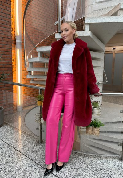 Abrigo de piel sintética burdeos sobre una camiseta blanca, combinado con pantalones de cuero rosa brillantes y tacones negros de puntera. Escalera de caracol al fondo.