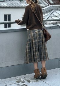 Pulover maro tricotat, fustă plisată în carouri în tonuri închise și ghete scurte din piele întoarsă de culoare cămilă. Include o geantă de umăr și un decor cu acoperiș acoperit de zăpadă.