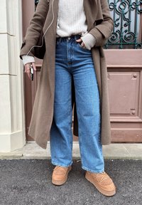Long manteau brun par-dessus un pull en maille crème, jean large bleu, et chaussures plateforme marron clair avec des accents texturés, debout sur un trottoir sombre.