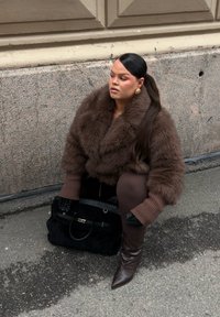 Brun pälsjacka i konstmaterial med bred krage och texturerade ärmar. Svart handväska med silverdetaljer. Mörkbruna figurnära byxor och spetsiga stövlar.