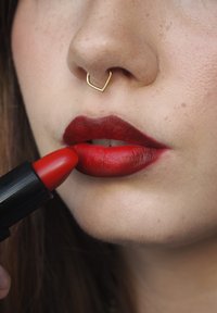Röd läppstift appliceras på fylliga läppar. Personen har en guld septumring och en slät hy med synliga fräknar.