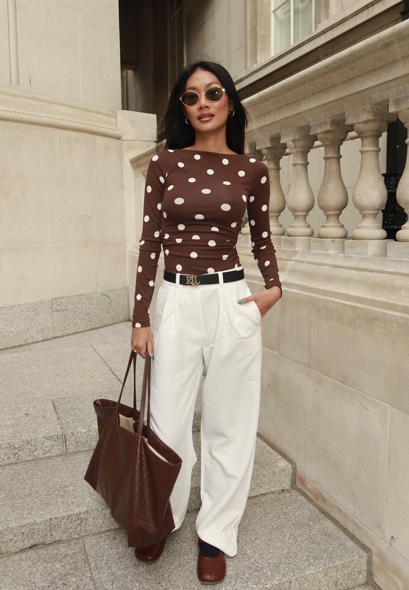 Femme portant un haut marron à pois, un pantalon blanc taille haute avec ceinture noire, des chaussures marron, des lunettes de soleil et tenant un grand sac cabas marron, debout sur des marches en pierre.