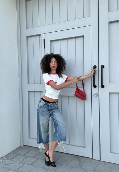 Camisa blanca con ribete rojo y diseño gráfico, jeans cortos en azul desteñido con dobladillo deshilachado, zapatos de tacón negros y un pequeño bolso rojo frente a una puerta gris.