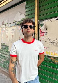 Maglietta bianca con dettagli rossi, con un piccolo logo Wrangler. Jeans blu e occhiali da sole, con uno sfondo urbano e strutturato.