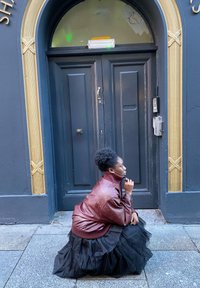 Bordowa skórzana kurtka na bluzce z wysokim kołnierzem, zestawiona z warstwową czarną tiulową spódnicą. W tle ciemne drzwi ze złotymi zdobieniami.