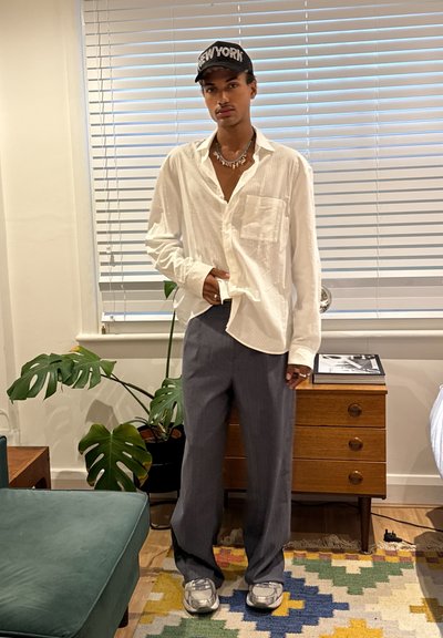 Camisa blanca de manga larga y botones con bolsillo; pantalones gris con pleats; zapatillas plateadas; gorra negra con el texto "NUEVA YORK"; ambiente interior.