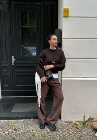 Frau in dunkelbraunem Pullover und weiten Hosen, die eine schwarze Handtasche mit einem weißen Schal hält, steht auf einer Kopfsteinpflasterstraße neben einer dunklen Tür und einer beige Wand.