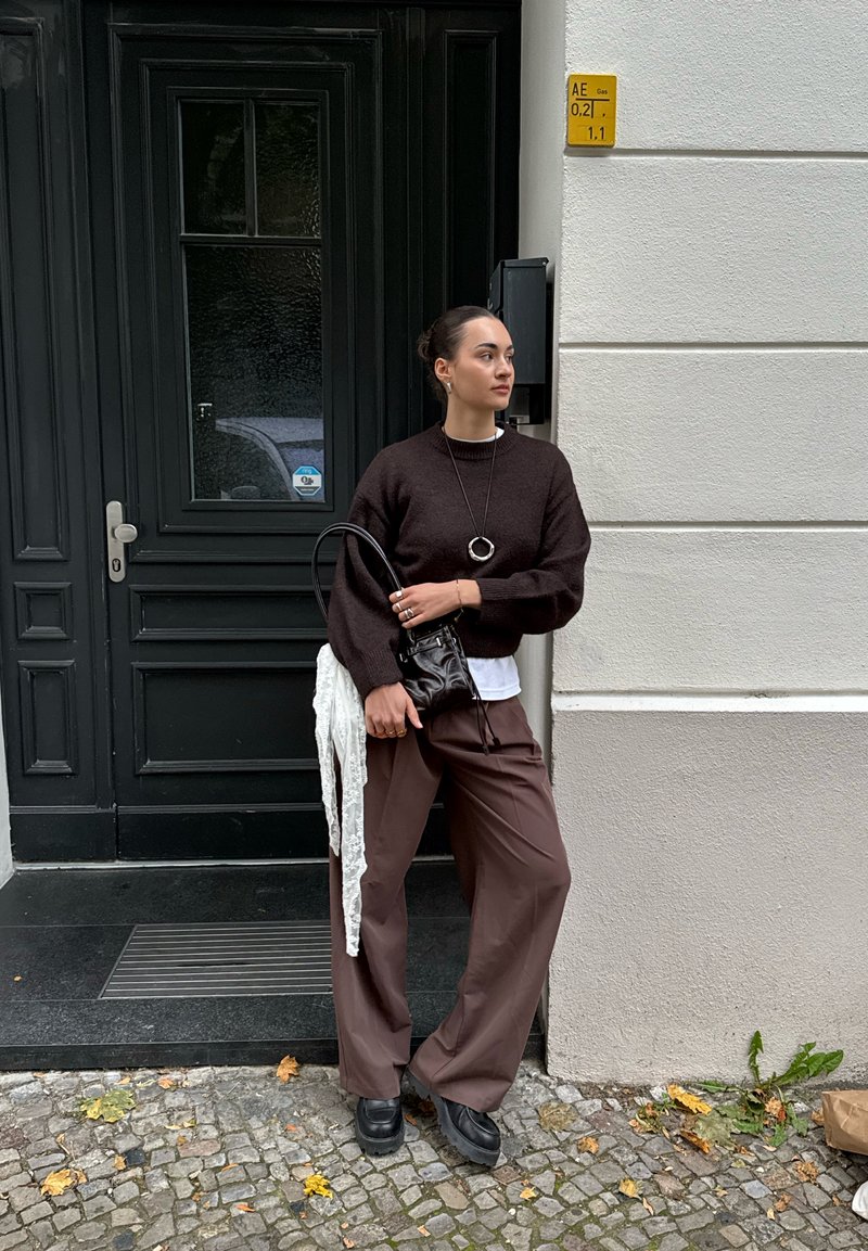 Femme portant un pull marron foncé et un pantalon large, tenant un sac à main noir avec une écharpe blanche, se tenant sur une rue en mode pavé près d'une porte sombre et d'un mur beige.