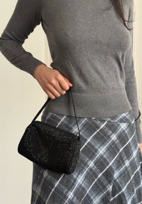 Personne tenant un petit sac à main noir à perles, portant un pull gris et une jupe à carreaux gris.