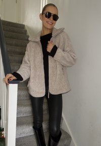 Veste en polaire beige avec un col montant et une fermeture éclair, associée à des leggings noirs et des bottes noires montant jusqu'aux genoux, sur fond d'un escalier.