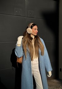 Femme aux longs cheveux portant un manteau bleu, des moufles blanches, un col roulé crème, un pantalon blanc et des cache-oreilles beiges, debout contre un mur sombre.