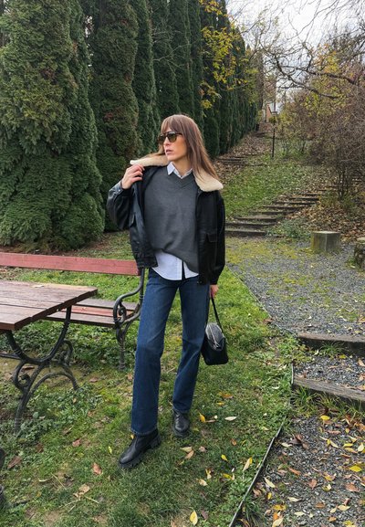 Chaqueta de cuero negra con cuello de borrego, suéter gris, camisa blanca, jeans acampanados azules, botas negras y pequeño bolso negro en un entorno natural.