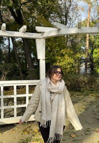 Beige nahkatakki, kerma huivi hapsuilla, mustat housut. Seisoo puistossa, taustalla vihreää kasvillisuutta ja pudonneita lehtiä.