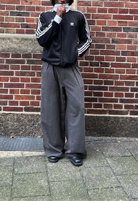 Černá bunda na zip s bílými pruhy, doplněná širokými šedými plisovanými kalhotami a černými botami. Stojící před zdi z cihel.