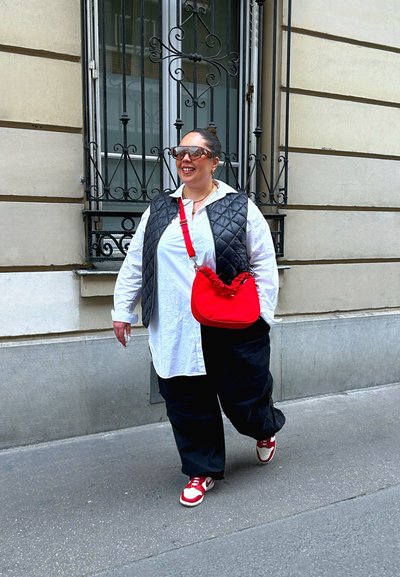 Chaleco de nailon negro acolchado sobre una camisa blanca, pantalones negros sueltos, zapatillas rojas y un bolso rojo con la parte superior fruncida. Entorno urbano.