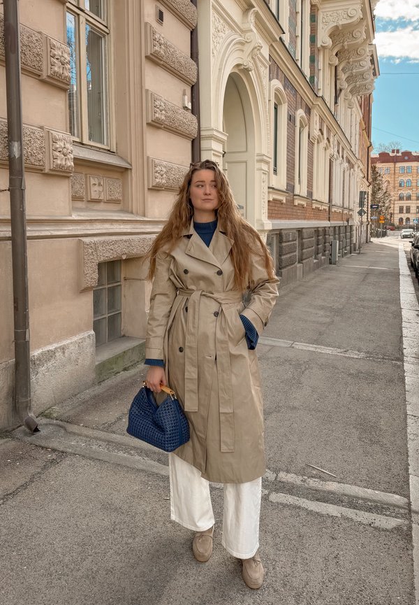 Femme aux longs cheveux portant un trench beige, un pantalon blanc et tenant un sac à main bleu, debout sur un trottoir en ville près d'un bâtiment orné.