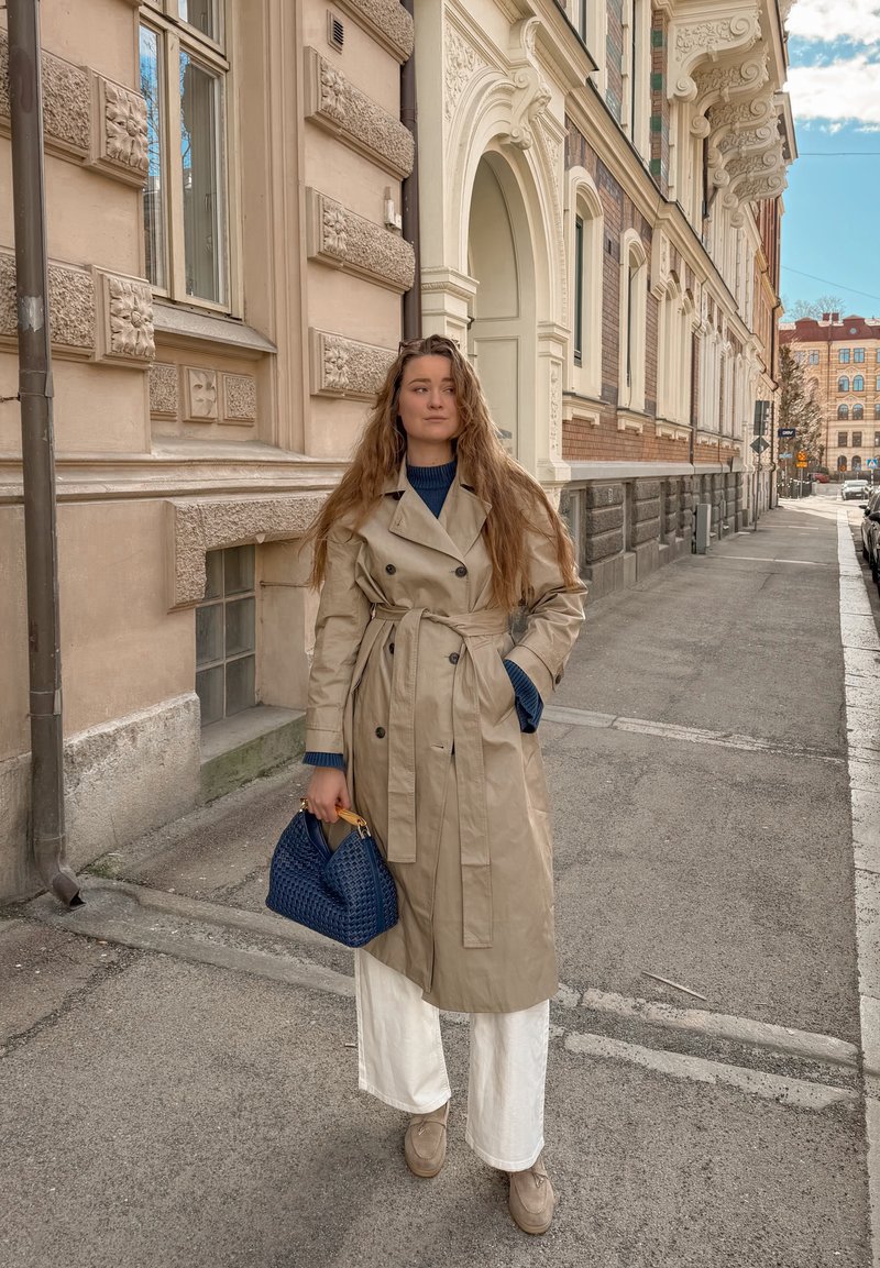 Femme aux longs cheveux portant un trench coat beige, un pantalon blanc et tenant un sac à main bleu, debout sur un trottoir en ville près d'un bâtiment orné.