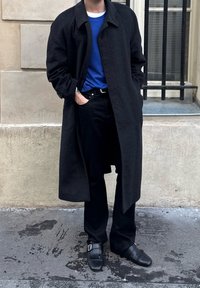 Άτομο που φοράει μακρύ μαύρο παλτό, μπλε πουκάμισο, μαύρο παντελόνι και μαύρα παπούτσια, στέκεται με το ένα χέρι στην τσέπη σε πεζοδρόμιο της πόλης.