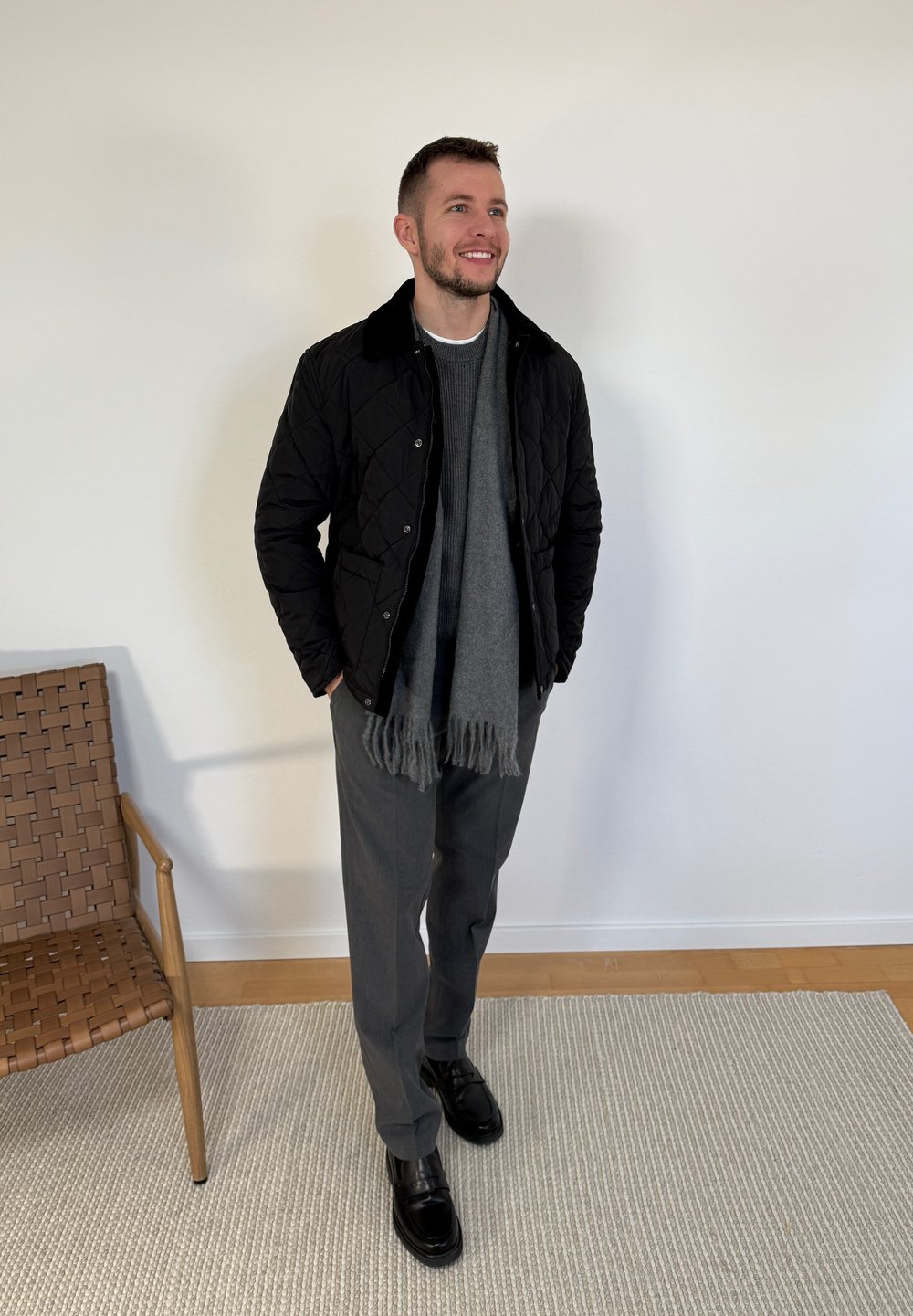 Homme debout à l'intérieur portant une veste matelassée noire, une écharpe grise, un pantalon gris et des chaussures noires à côté d'une chaise en osier sur un tapis.