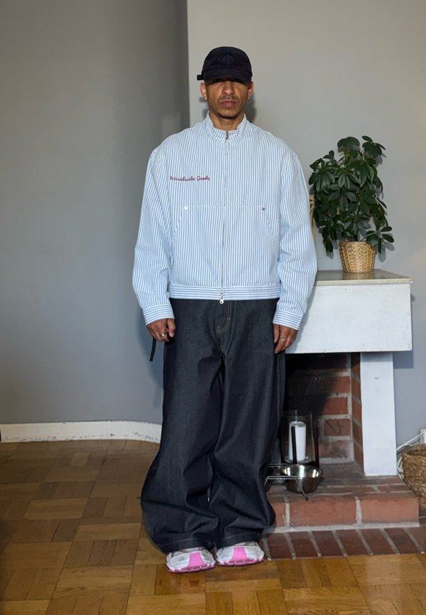 Homme portant une casquette noire, une veste zippée blanche à rayures, un pantalon large foncé et des baskets blanches avec des semelles roses, debout à l'intérieur près d'une petite cheminée blanche.