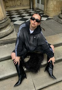 Hanorac negru cu fermoar și dungi albastre, pantaloni scurți din denim și cizme negre elegante cu vârful ascuțit. Model așezat pe trepte de piatră, ținând o pătură neagră texturată.