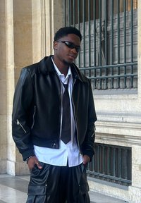 Black leather bomber jacket with zipper details, worn over a white shirt and dark tie, paired with black leather cargo pants. Sunglasses worn.