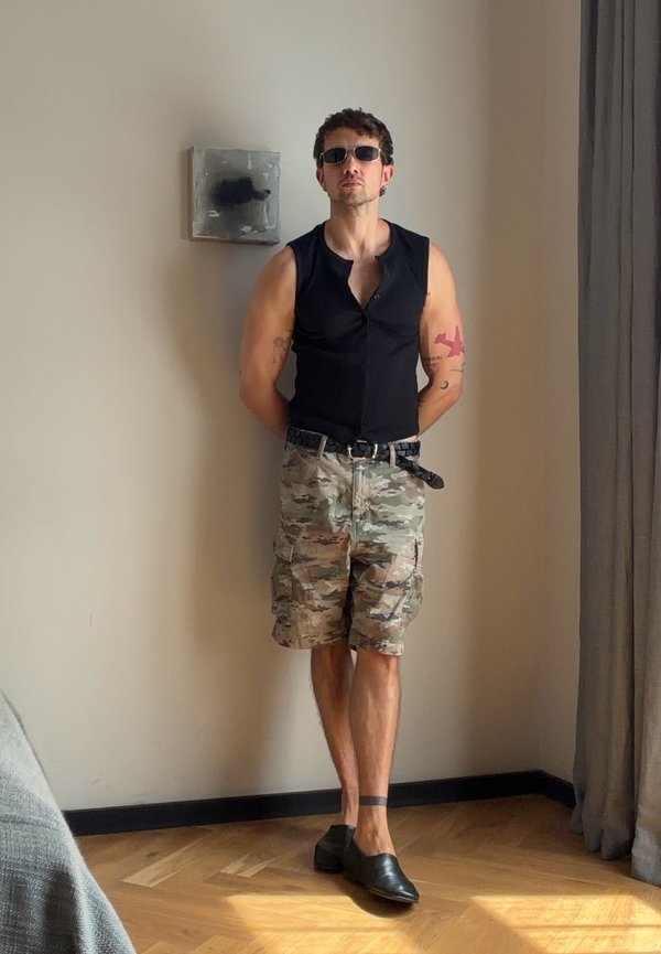 Man wearing sunglasses, sleeveless black shirt, camo shorts, and black shoes stands against plain wall in sunlit room with wooden floor.