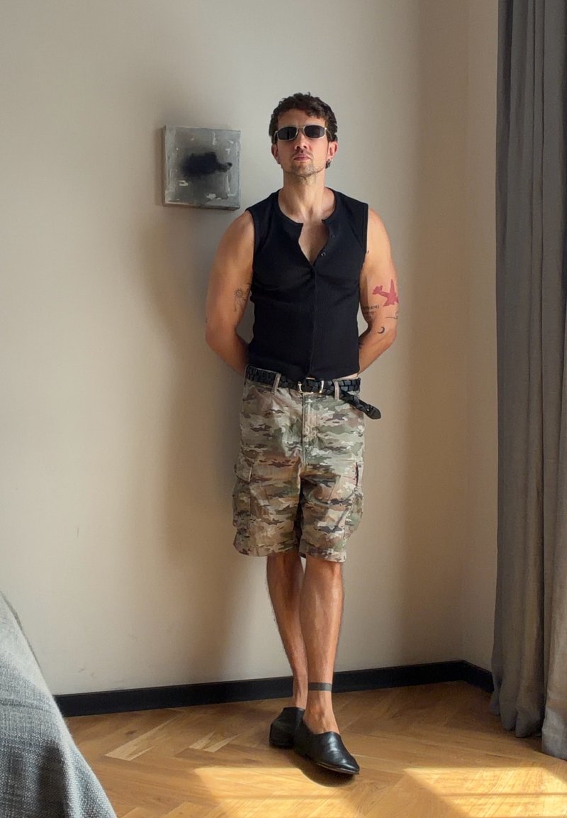 Homme portant des lunettes de soleil, un débardeur noir, un short camouflage et des chaussures noires se tient contre un mur uni dans une pièce ensoleillée avec un sol en bois.