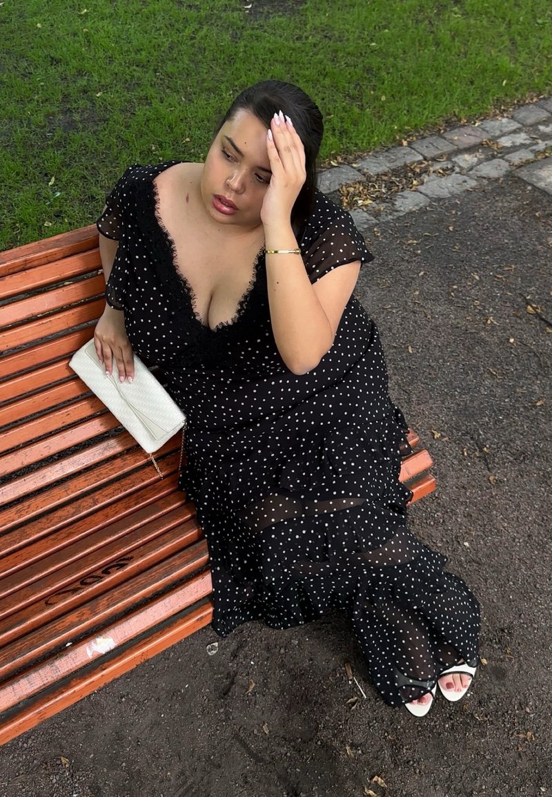 Femme en robe noire à pois, assise sur un banc en bois à l'extérieur, tenant une pochette blanche et touchant son front avec une expression pensive.