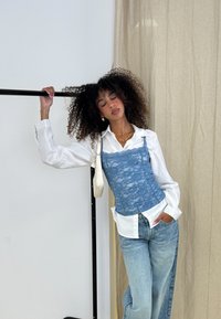Lyseblå spids top lagdelt over en hvid skjorte med knapper, parret med løse lyse denimjeans. Modellen står foran en neutral baggrund.