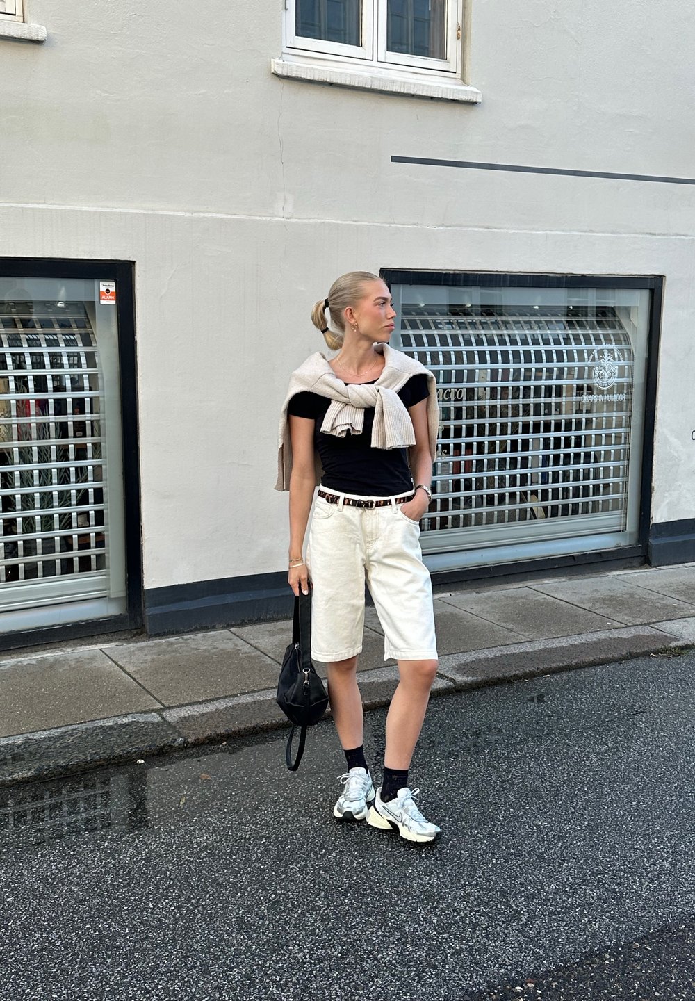 Μαύρο κοντομάνικο μπλουζάκι, άσπρα σορτς bermuda, μπεζ πουλόβερ δεμένο γύρω από τους ώμους, μαύρη τσάντα και ασημένια αθλητικά παπούτσια με ανακλαστικά στοιχεία σε βρεγμένο δρόμο.