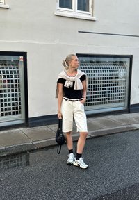 Μαύρο κοντομάνικο μπλουζάκι, άσπρα σορτς bermuda, μπεζ πουλόβερ δεμένο γύρω από τους ώμους, μαύρη τσάντα και ασημένια αθλητικά παπούτσια με ανακλαστικά στοιχεία σε βρεγμένο δρόμο.