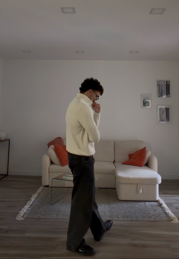Man in witte trui en zwarte broek loopt bedachtzaam in een minimalistische woonkamer met beige bank en oranje kussens.