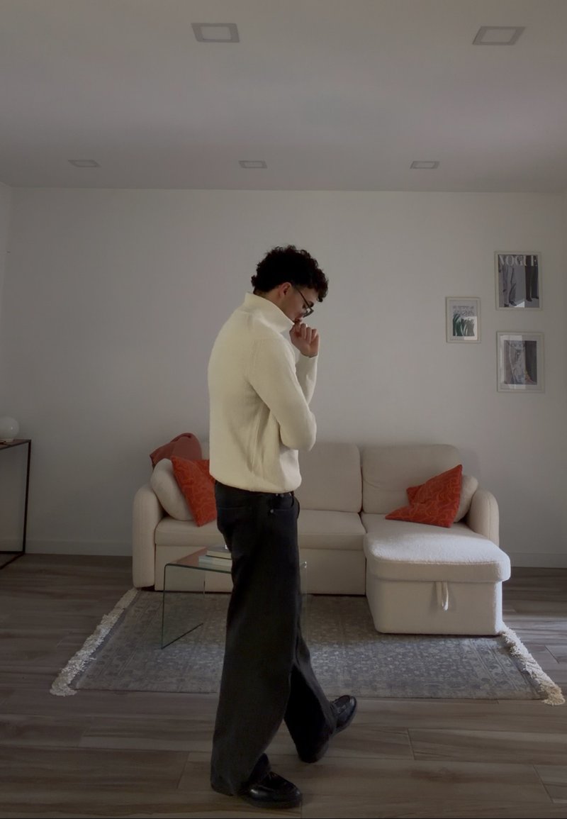 Mand iført hvid sweater og sorte bukser går eftertænksomt i et minimalistisk stue med beige sofa og orange puder.