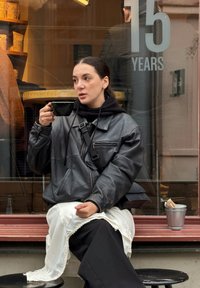 Veste en cuir noire à coupe décontractée. Vêtement blanc orné de dentelle en dessous. Tasse noire à la main. Seau argenté avec des serviettes à proximité.