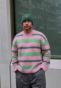 Suéter de rayas gruesas en rosa, verde y gris, hecho de un punto texturizado. Combinado con un gorro verde que presenta un parche de cuero marrón.