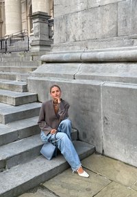 Grijze oversized blazer, lichtblauwe ripped jeans, witte puntige flats en een blauwe handtas, zittend op stenen trappen tegen een grijze muur.