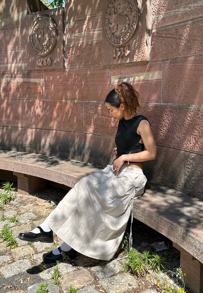 Top negro sin mangas, falda plisada beige clara con lazos ajustables y zapatos negros con calcetines blancos. Sentada en un banco de piedra.