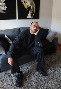 Prillidega ja habemega mees, kes kannab tumedaid riideid, istub elutoas hallil diivanil, millel on seinal abstraktsed näomaalid.