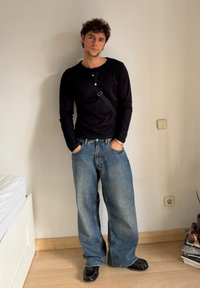 Mustmust kaelusega must särk, laiad sinised teksapüksid, mustad kingad, seistakse heleda seina vastu. Vabaaja riietus lahedas istuvuses.