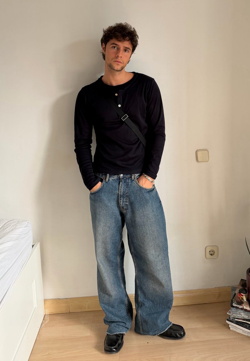 Czarna koszula z długim rękawem z guzikami, luźne niebieskie dżinsy, czarne buty, stojąca przy jasnej ścianie. Swobodny strój w luźnym fasonie.