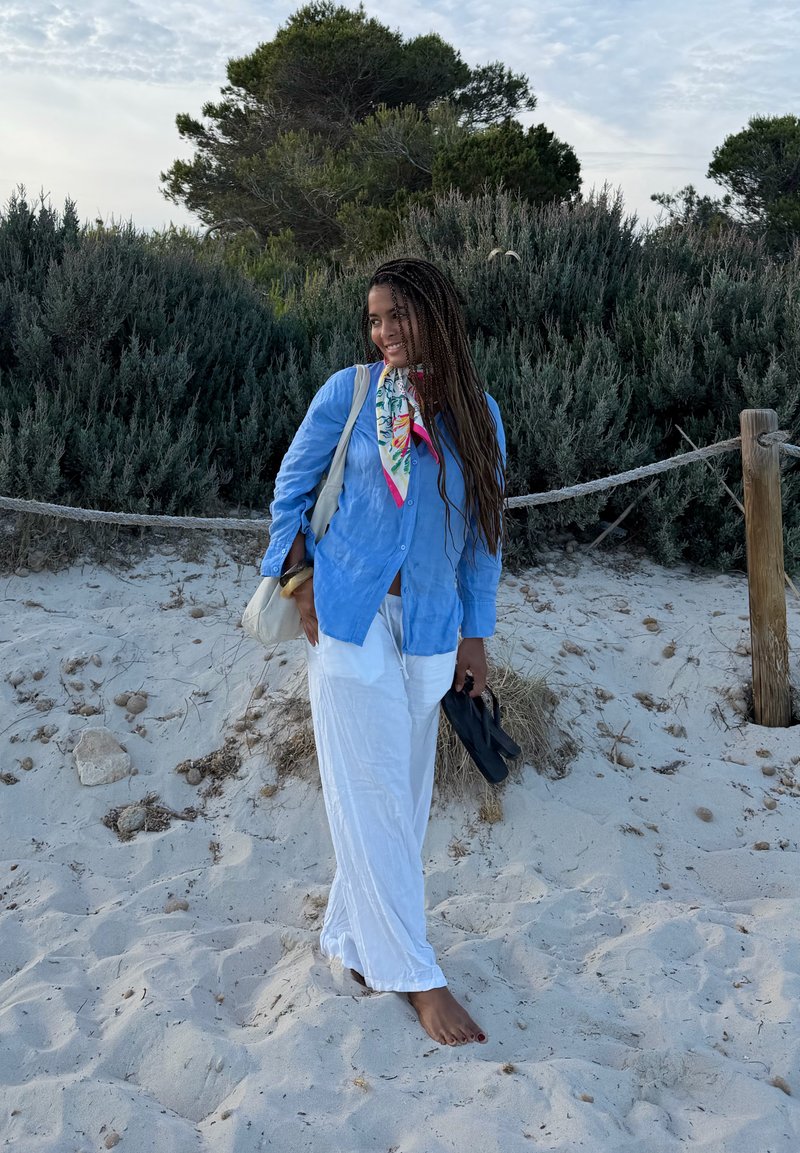 Femme avec de longues tresses, portant une chemise bleue et un pantalon blanc, debout pieds nus sur une plage de sable près de la verdure tenant des sandales noires.