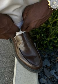 Καφέ δερμάτινες loafers με λεία υφή, που διαθέτουν μια διακοσμητική ζώνη κατά μήκος του επάνω μέρους. Φοριούνται με άσπρες κάλτσες και ανοιχτόχρωμο παντελόνι.