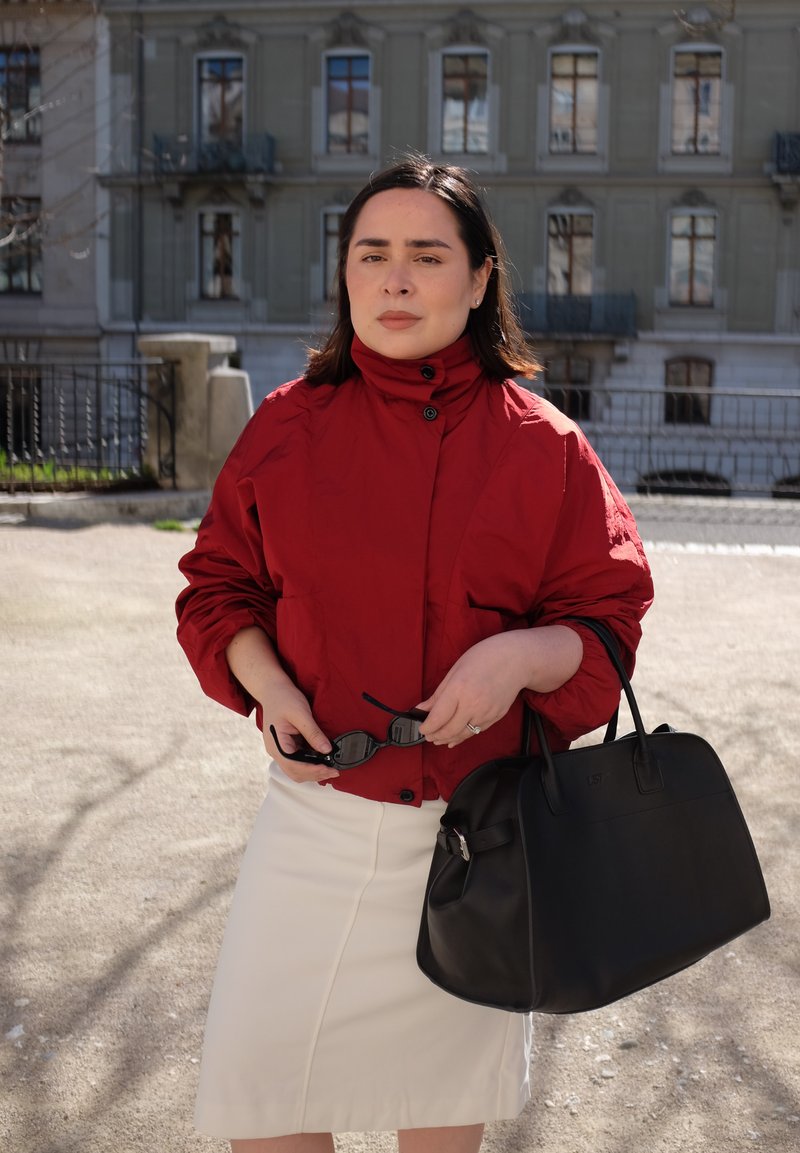 Femme debout à l'extérieur avec une veste rouge et une jupe blanche, tenant un sac à main noir et des lunettes de soleil avec un bâtiment à plusieurs étages en arrière-plan.