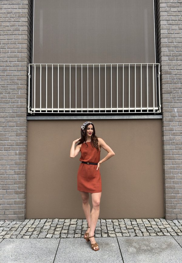 Jeune femme en robe couleur rouille et sandales posant sur un trottoir pavé devant un mur beige avec un petit balcon au-dessus.