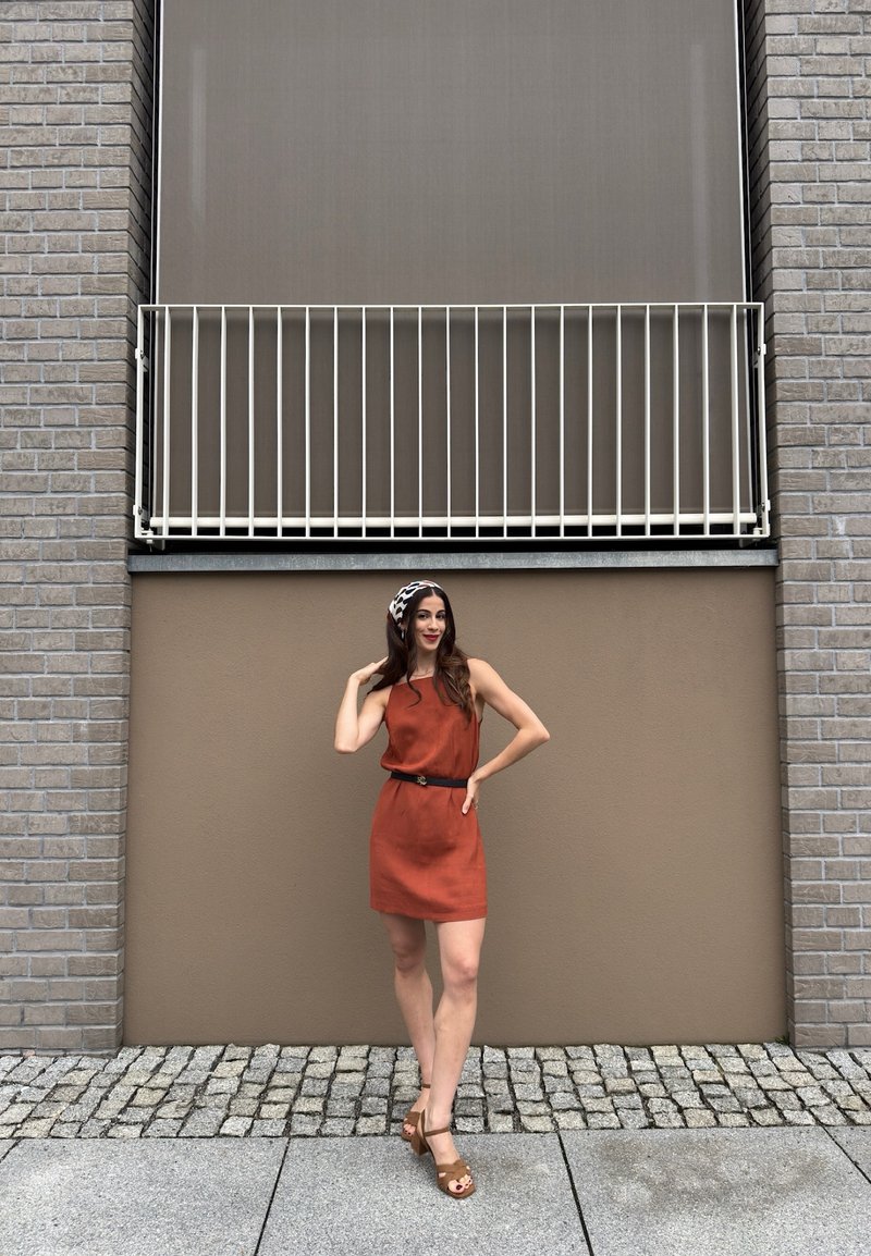 Jeune femme en robe couleur rouille et sandales posant sur un trottoir pavé devant un mur beige avec un petit balcon au-dessus.