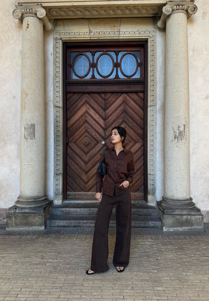 Femme en tenue marron foncé et sandales noires debout sur des marches en pierre entre de grandes colonnes devant une porte en bois ornée.
