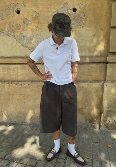 Camisa polo blanca, pantalones cortos a rayas oscuros, calcetines blancos, zapatos marrones sin cordones con detalles. Gorra de camuflaje, tatuajes visibles en los brazos.