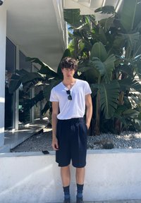 Maglietta bianca a V, pantaloni corti a pieghe blu navy, calze scure e scarpe nere. In piedi contro una parete bianca testurizzata con vegetazione sullo sfondo.