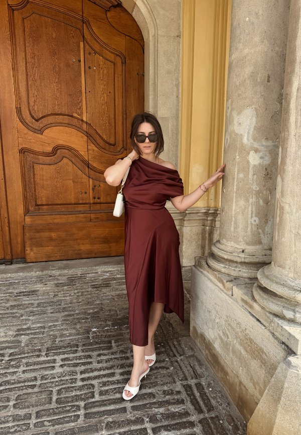 Žena u bordo haljini bez ramena i bijelim sandalama pozira kraj kamenih stupova i velikih ukrašenih drvenih vrata.